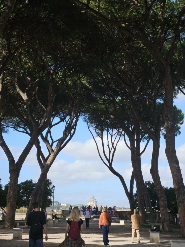 View of St Peter’s Basilica framed by trees in the Orange Garden on Aventine Hill with people walking.